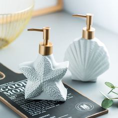 two white soap dispensers sitting on top of a book next to a plant