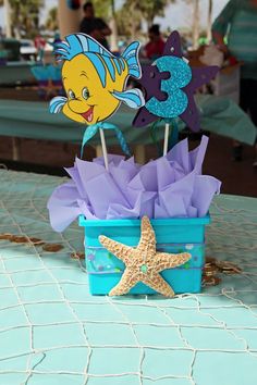 a blue bucket filled with purple tissue paper flowers and two starfish toppers sitting next to each other