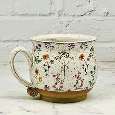 a white and brown coffee cup with flowers on it sitting on a table next to a brick wall