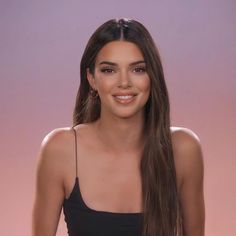 a woman with long brown hair wearing a black top and smiling at the camera while standing in front of a pink background