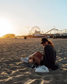 a woman sitting on top of a sandy beach next to a ferris wheel at sunset