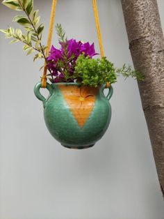 a green vase with purple flowers in it hanging from a tree