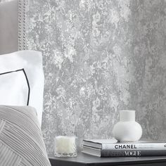 a nightstand with two books on it next to a white vase and lamp in front of a gray wall