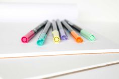 five markers are lined up on top of a piece of white paper with orange, green, and blue marker pens