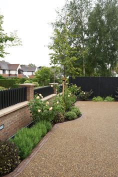 a paved driveway with flowers and trees in the back ground, surrounded by black fence