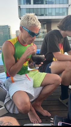 a man sitting on top of a wooden floor next to another person wearing sunglasses and eating food