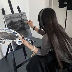 a woman sitting at a table painting with headphones on her ears and holding a paintbrush