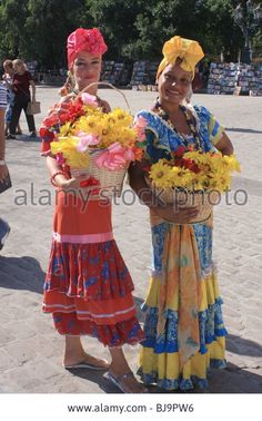 two women dressed in colorful clothing holding flowers and baskets on their heads, posing for the camera