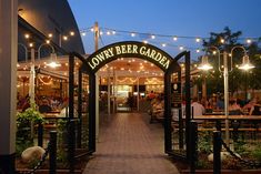 an entrance to a beer garden with lights strung over it and people sitting at tables in the background