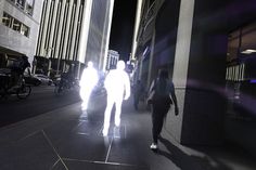 people walking down the street at night with their shadows on the ground and buildings in the background