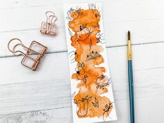 an orange and black watercolored bookmark next to some office supplies