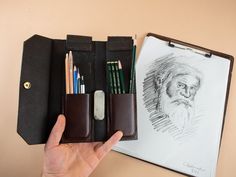 a hand holding a pen and pencil case next to a drawing book with an old man's face on it