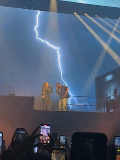 two people standing on top of a stage with cell phones in front of them and lightning behind them