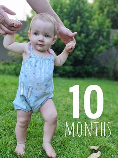 a baby standing in the grass with its hands on it's mother's arm