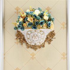 a decorative wall hanging with flowers in a vase on top of the wall and an ornate gold frame around it