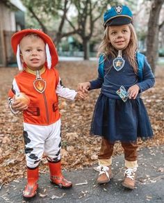 two young children dressed in costumes standing next to each other on a sidewalk with leaves all over the ground