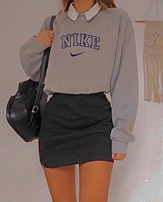 a woman in grey sweater and black skirt holding a handbag while standing next to a wall