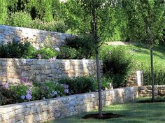 a stone wall with flowers growing on it and trees in the foreground, next to a grassy field