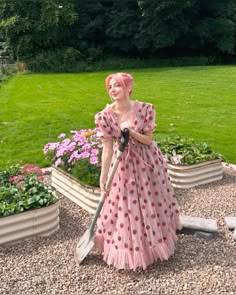 a woman in a pink dress is holding a shovel and standing next to some flowers