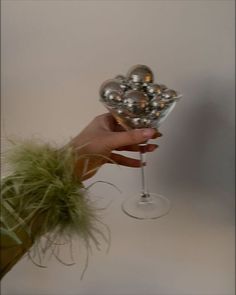 a hand holding a wine glass filled with silver balls and greenery in front of a white wall