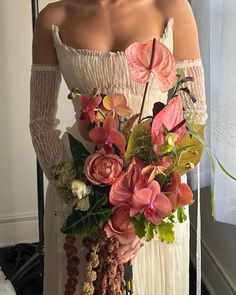 a woman in white dress holding a bouquet of flowers and greenery next to a window