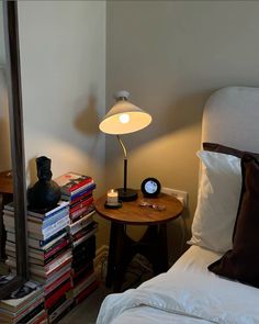 a bed with white sheets and pillows next to a night stand, lamp and books