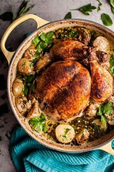 a chicken dish with potatoes and herbs in a yellow pan on a blue towel next to green leaves