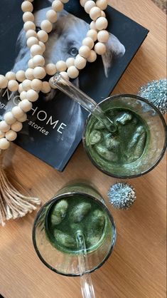 two glasses filled with green liquid on top of a wooden table next to a book