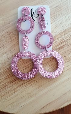 pink glitter earrings with circles and rings on top of a wooden table next to a card