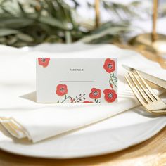 a place card on a plate with gold forks and napkins next to greenery