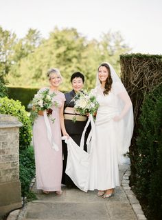 three women standing next to each other in front of a stone wall with greenery