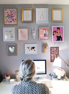 a woman sitting in front of a computer on top of a desk next to a wall covered with pictures