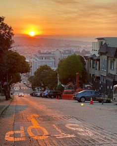 the sun is setting over a city street with cars parked on both sides and buildings in the background