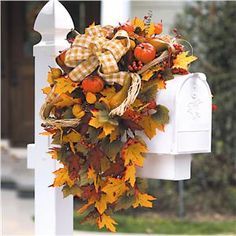 a wreath is hanging on the mailbox in front of a white fence with autumn leaves and pumpkins