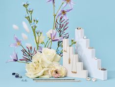 an arrangement of flowers and jewelry sitting on a blue surface next to a stack of white boxes