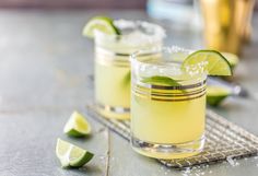 two glasses filled with lemonade and limes on top of a metal tray next to some sliced limes