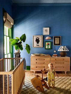 a baby standing next to a wooden rocking horse in a room with blue walls and pictures on the wall