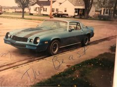 an old photo of a blue car parked on the side of the road in front of some houses