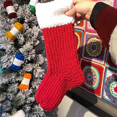 a person holding a knitted stocking next to a christmas tree with decorations on it