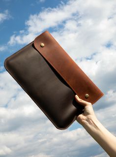 a hand holding a brown leather case in front of a blue sky with white clouds