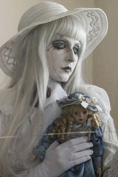 a woman with white hair and makeup holding a doll in her hands while wearing a hat