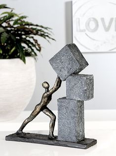 a statue of a person pushing a block with one hand on top of it, in front of a potted plant