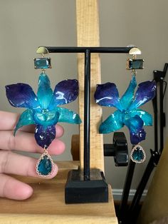 These stunning earrings feature real blue orchids delicately preserved in resin, creating a unique and eye-catching piece of jewelry. The orchids are complemented by gold-plated half moon studs and elegant blue glass charms, adding a touch of sophistication to the design. **Details - Real blue and purple orchids preserved in resin - Gold-plated half moon studs - Blue glass charms - Drop length: 3-4 inches Each pair of earrings is handmade with care, making them a perfect gift for nature lovers or anyone who appreciates beautifully crafted jewelry. Add a pop of color and elegance to your outfit with these exquisite orchid earrings! The colors and shapes of each orchid might be slightly different as they are made of real orchid flowers.This is a precious item that deserves your utmost attent Elegant Blue Resin Earrings, Blue Resin Jewelry For Parties, Blue Resin Jewelry For Party, Blue Flower-shaped Resin Earrings, Blue Flower Resin Jewelry, Blue And Purple Orchids, Orchid Earrings, Orchid Flowers, Blue Orchids