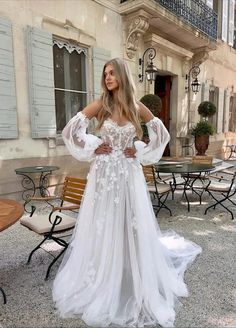 a woman standing in front of a building wearing a white wedding dress with sheer sleeves