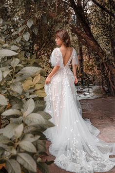 a woman standing in front of some trees wearing a wedding dress with flowers on it