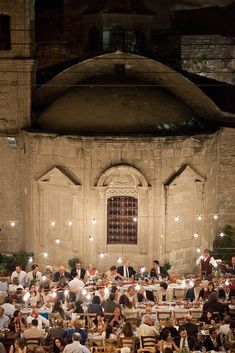 a large group of people sitting at tables in front of a building with lights on it