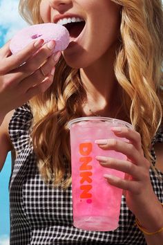 a woman eating a doughnut while holding a pink drink