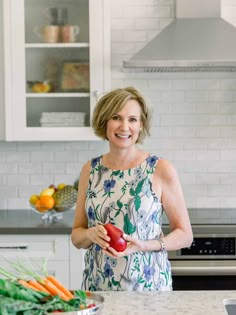 a woman standing in a kitchen holding a red apple and an orange on the counter