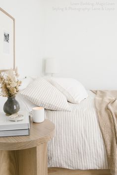 a bed with white sheets and pillows next to a table with a vase on it