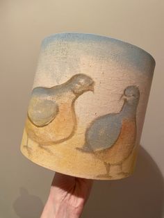 a hand holding a lamp shade with two birds painted on the lampshade,